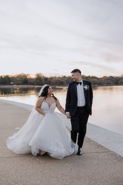 Couple in wedding dress and suit hold hands walking along side the water.