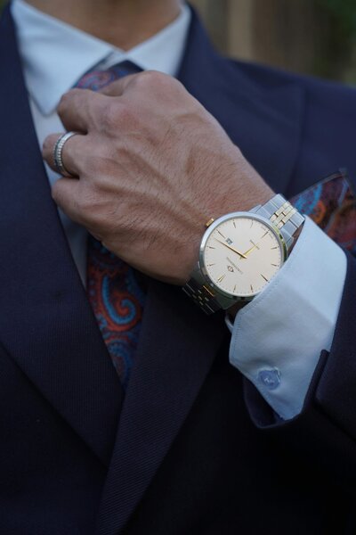 A close-up of a man adjusting his navy blue tie, with a focus on his gold and silver wristwatch and pocket square, wearing a crisp white shirt under a formal suit.