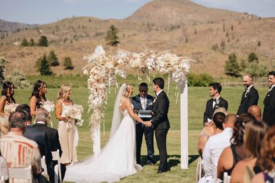 Couple Under Floral Ceremony Backdrop - Mikayla & Mario | Harmony Meadows Wedding - Lake Chelan Wedding