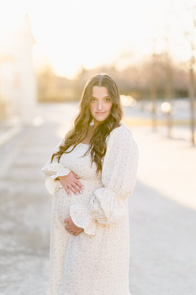 A pregnant woman wearing a white dress looks directly into the camera.