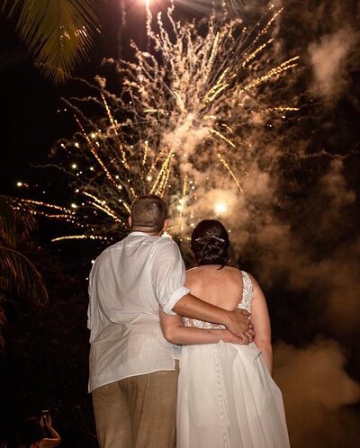 A bride and groom embrace while watching a stunning fireworks display at their destination wedding reception, celebrating their special day under the night sky.