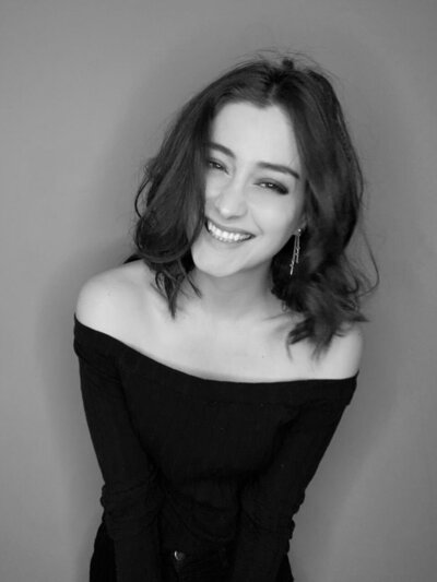 Black and white photo of woman with short, curly  brown curly hair  smiling at camera wearing a black off the shoulder top.