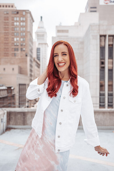 woman with red hair wearing a tie dye dress and white jacket standing on a rooftop in downtown Cincinnati Ohio and smiling at the camera