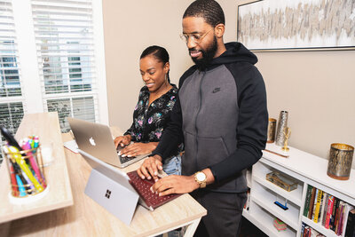 A man and woman working side by side at a standing desk, both using laptops. The man, dressed in a hoodie, types on a Surface laptop, while the woman, dressed in a floral blouse, works on a MacBook. They are focused and collaborating in a home office environment