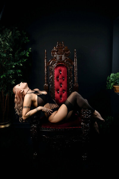 A woman in black lingerie and fishnet stockings reclines gracefully on an ornate red velvet chair. The setting is dark, with lush green plants.