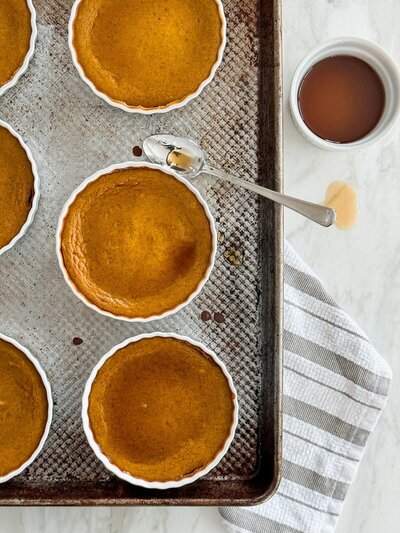 Individual dairy-free pumpkin custards on a rimmed baking sheet, next to a bowl of maple syrup with a spoon.