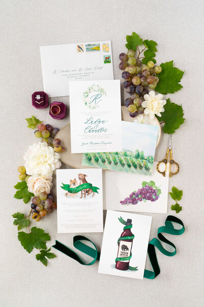 Elegant vineyard-themed wedding invitation suite by E Stokes Creations, featuring watercolor illustrations of grapes, a vineyard, and a wine bottle, accented with green and purple details. The suite includes invitations, RSVP cards, and envelopes, adorned with floral elements, vintage scissors, and wedding rings, perfect for a vineyard or winery wedding.