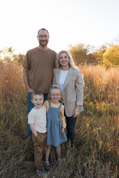 A smiling family of four poses in a field, with warm autumn colors creating a beautiful backdrop.