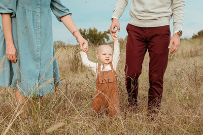 A sweet family moment in a golden field near Charlotte, with parents holding their toddler’s hand during a photography session