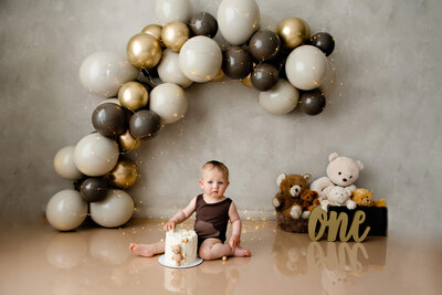 1 year old baby sitting in front of a balloon arch with brown tones for his teddy bear cake smash photography session in Burlington, Ontario.