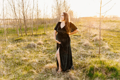 stunning mother of 3 holding her baby bump and looking into the field as the sun sets behind her
