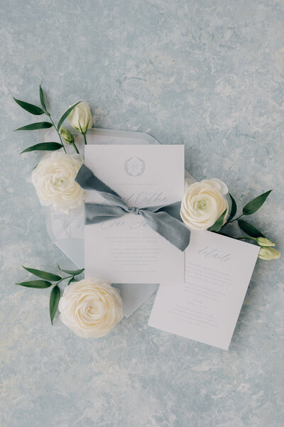 A blue and white invitation with blue ribbon invites guests to The Acreage - a wedding venue near Omaha NE. Photo by Anna Brace, an Omaha Wedding Photographer