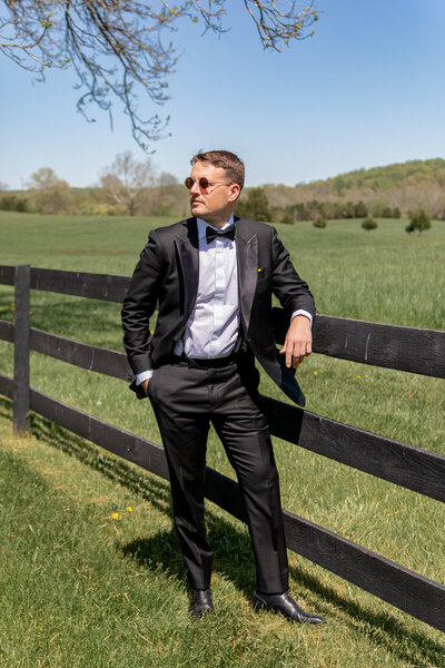 A groom poses for a portrait on his wedding day. Leaning against a black wooden fence in a grass field, he's looking to the side in a candid way.