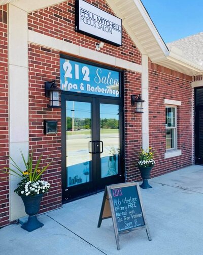 The front entrance of 212 Salon, Spa, & Barbershop, featuring a brick exterior, a glass door with the salon's name, and a welcoming chalkboard sign outside.