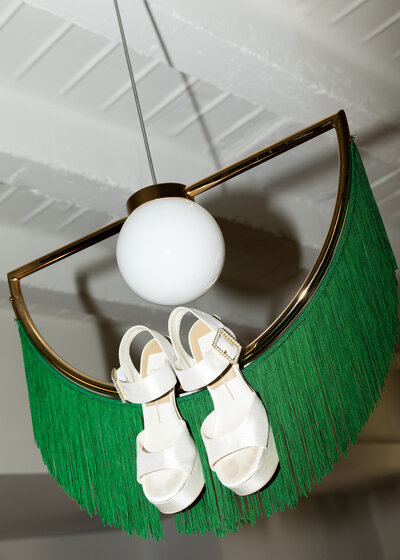 bridal shoes hanging from a mid century modern light fixture with green fringe photographed as an editorial wedding detail