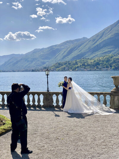 A couple standing along a railing in front of the water while a photographer takes a picture of them.