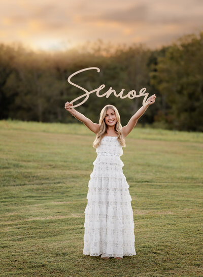 Senior photo session in an open field of girl holding up Senior wooden sign. Atlanta Photographer, jenni Nikki Photography, took the photo.