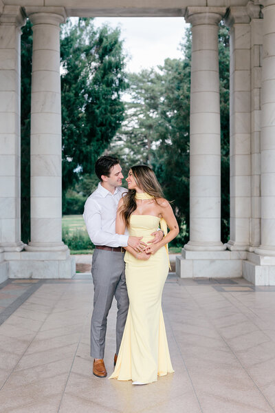 couple elegantily dressed, posing for a photo and looking at each other on Cheesman Park in Denver. Behind the the white columns of the building.