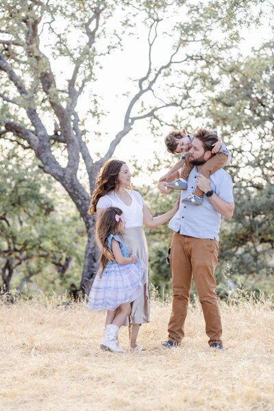 Candid family moment during San Jose golden hour session: toddler son riding on father's shoulders while young daughter hugs mother