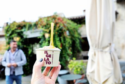 Hand holding a cup of ice cream in front of a greenery-draped building.