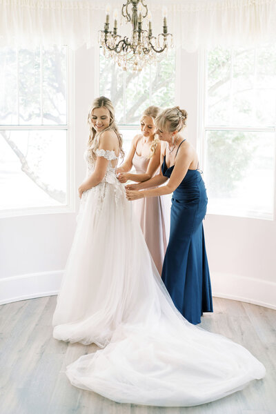 Bride in a white wedding dress being helped with the back of her dress by two women inside a room with a chandelier and large windows, organized meticulously by a renowned Calgary wedding planning team.