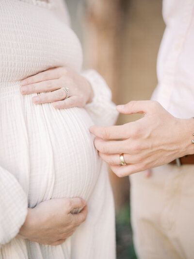 Husband's hand grazing wife's pregnant belly as she holds the top and bottom of her belly.