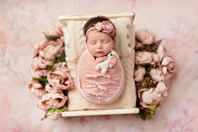 Newborn baby girl wrapped in pink, sleeping on a small cream bed surrounded by pink florals and holding a tiny white teddy bear.