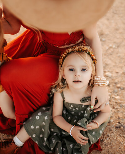 Little girls looking up at the camera in a green floral dress with a flower crown sitting next to her mom in a red dress