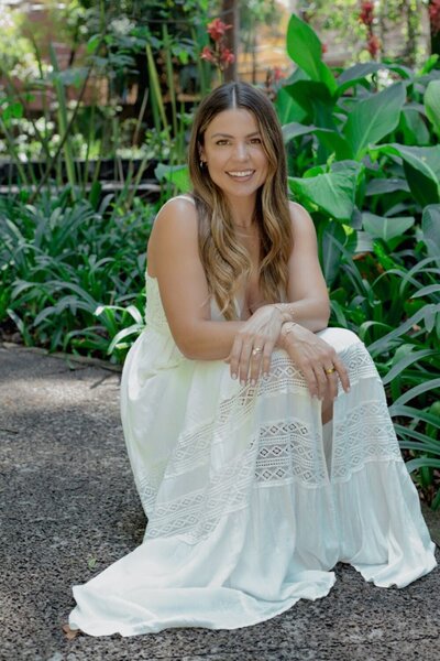 Natalie Bernacchi smiling and seated in a garden, wearing a white lace dress surrounded by lush green plants