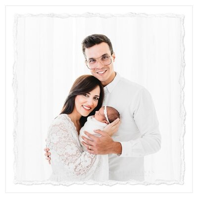 The photo shows a proud mom and dad holding their newborn baby, dressed in all white. The setting is a pristine all-white studio, creating a clean and pure atmosphere. The photographer, Bri Sullivan, has captured the emotional bond between the parents and their child, highlighting the joy of welcoming a new addition to the family.