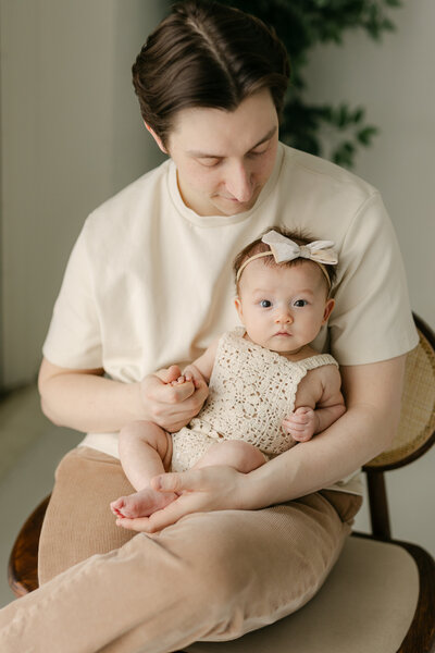 An infant girl in a knit onesie and bow headband sits in dad's lap