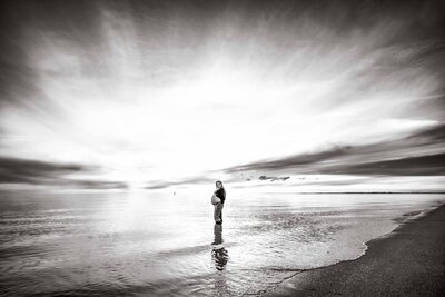 Black and white image of a pregnant mother in jeans and a jumper showing her bump in the water at the beach at dusk