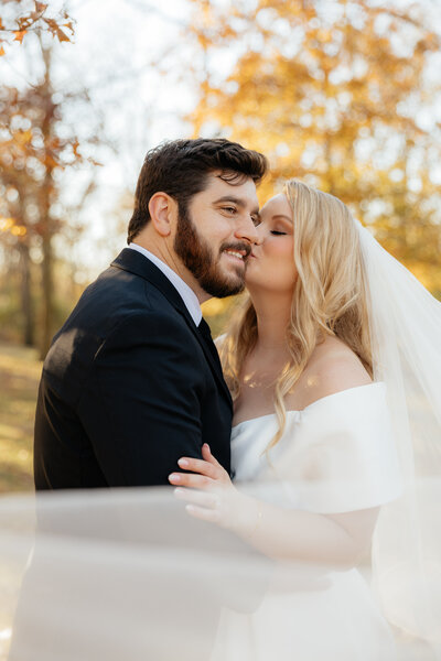 The bride kissing the groom on the cheek with their veil draped in front of them.