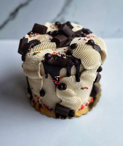Photo shows a small cutting cake with a slightly tan coffee frosting. There is some white strips of squiggly frosting with ridges randomly over the cake. There is also chocolate drip, chocolate covered coffee beans, chocolate candies, and rainbow sprinkles scattered throughout the cake as decorations.