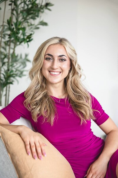 Smiling woman in a bold fuchsia dress sits confidently on a couch with soft, neutral decor and a green plant in the background. Captured during a personal branding photoshoot, this polished and approachable image conveys leadership, professionalism, and vibrant energy.
