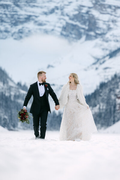 Bride and groom standing on the frozen surface of Lake Louise in winter with snow covered surroundings and Victoria Glacier in the background captured as a scenic wedding portrait