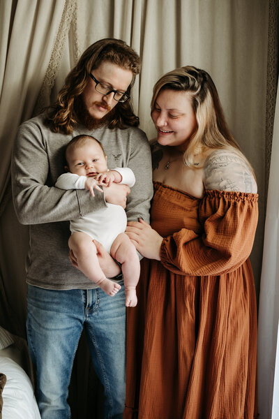 Smiling mom in rust dress looking at newborn baby boy wearing white while dad is holding baby in gray and jeans in Annapolis Maryland.