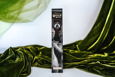 High priestess incense in its packaging — A long, dark, and sleek envelope with Roman-style statues of nymphs. The top shows the Lowborn Goods logo, below that is the high priestess motif with a haloed woman whose face is obscured by smoke coming from her mouth. The center of the packaging reads "high priestess" in gothic font. Below the name lists "Bergamot + Hemp + Vetiver." At the bottom, it reads "20 Hand-dipped charcoal incense sticks" and "lowborngoods.com" Behind the packaging is green velvet and green tulle on a white background.