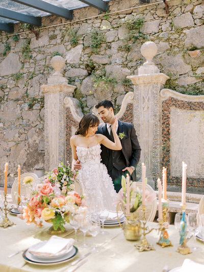 bride and groom embrace at reception at Casa dos Penedos in Sintra, Portugal with orange and pink flowers, peacock candlesticks, and rocky architecture in greenhouse room