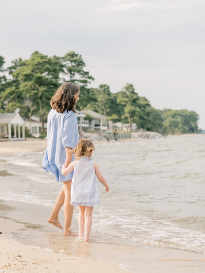 Mom wearing a blue dress holding daughters hand while going into Virginia Beach waters. 