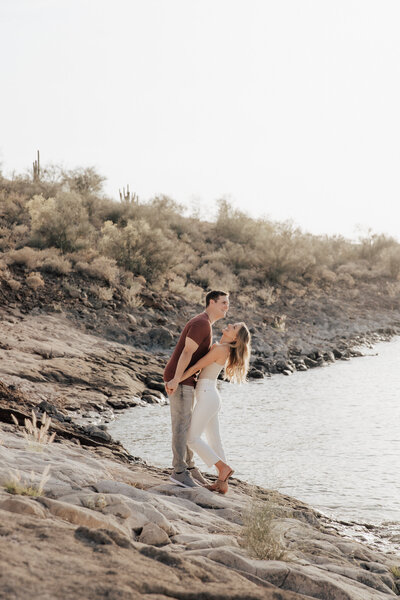 Couple holding hands and laughing on white rocks at Lake Pleasant in Arizona during golden hour engagement photos.