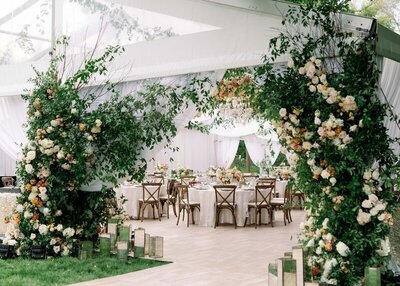 peering into wedding reception set up in minneapolis minnesota with tables and chairs and greenery with flowers creating arched entry into white tent