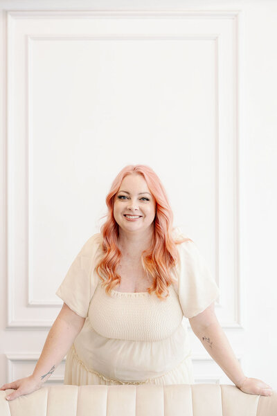 The Head Hair and Makeup Stylist leaning against back of white couch with curled pink hair