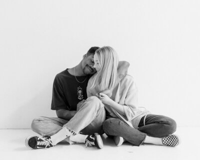 Black and white image of a couple sitting on the ground leaning against each other in a white studio