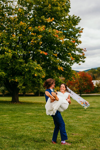 Groom holding bride while spinning her around with her veil flying in the wind under a big tree 