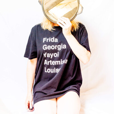 Henrie Richer wearing a shirt mentioning Frida, Georgia, Yayoi, Artemisia, Louise and holding a mirror