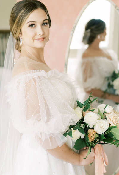 Portrait of a smiling bride in a white wedding gown holding a bouquet of white and blush roses in front of a mirror