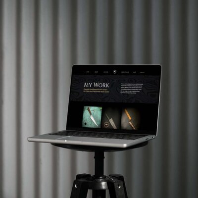 A laptop displaying a website for handcrafted kitchen knives, titled 'My Work' with images of various knives on a dark background. The laptop is set on a minimalist black stand with a curtain backdrop.