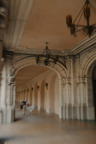 A perspective shot looking down a long outdoor hallway or loggia at a historic estate. The scene features a series of stone arches, hanging rustic chandeliers, and warm evening light.