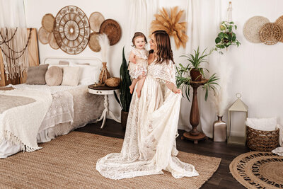 Mom is holding toddler daughter while swaying back and forth in a lace dress in a studio.